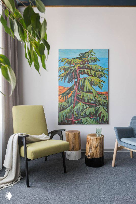 Уютный уголок с зелёным креслом и маленьким столиком, на стене висит крупная живописная картина с изображением дерева и яркими красками.