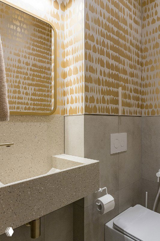 Угол гостевого санузла: золотой орнамент на стенах, панель инсталляции с кнопкой слива и держатель для туалетной бумаги на фоне фактурной столешницы.