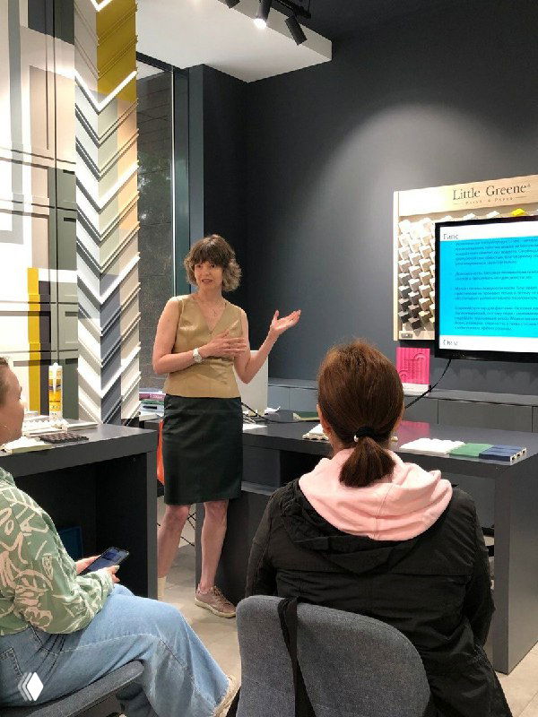 Лектор рассказывает в салоне-выставочном пространстве: женщина у стенда с образцами плитки и внимательные слушатели в зале на экскурсии с учениками.