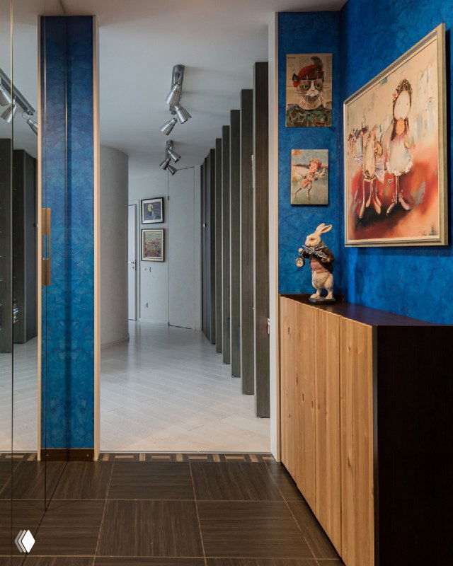 Длинный коридор с синими стенами и картинами; вертикальные колонны и деревянный шкаф показывают вариант откосов из дерева.