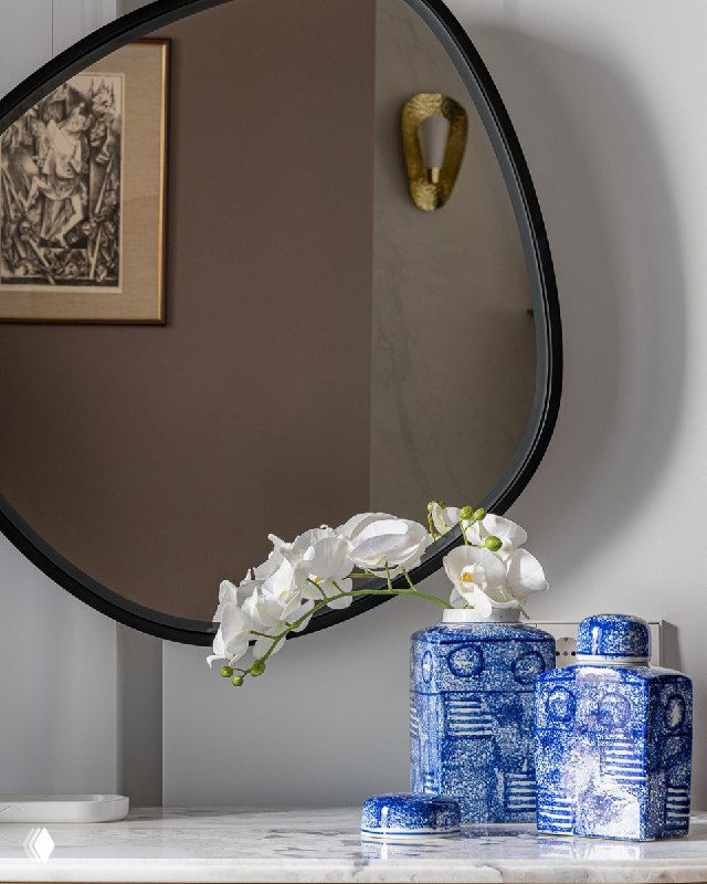 Крупный план круглого зеркала в тонкой черной раме над комодом с синими декоративными вазами и белым орхидеей, спокойный интерьерный свет.
