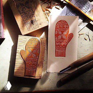 Резная матрица для линогравюры в виде варежки на рабочем столе: блок линолеума с аккуратной резьбой, готовый бумажный оттиск и инструменты для резки рядом.