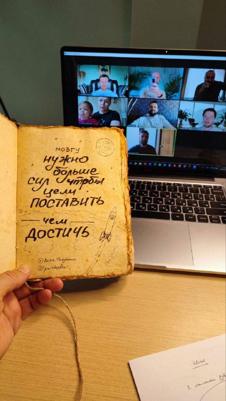 Открытая страница тетради с рукописной заметкой о целях и достижениях на фоне ноутбука и бумаги на столе.