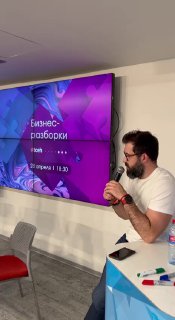 Ведущий говорит у большого экрана с слайдом «Бизнес‑разборки»: кадр с мероприятия, спикер с микрофоном объясняет кейс аудитории.