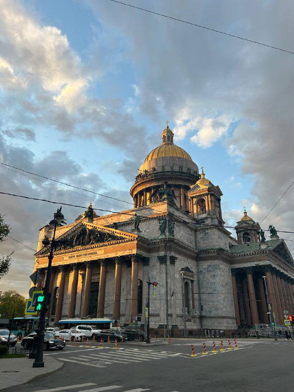 Исаакиевский собор в Санкт-Петербурге на фоне вечернего неба: купол в тёплом свете, улица, пешеходы и трамвайные провода в кадре.
