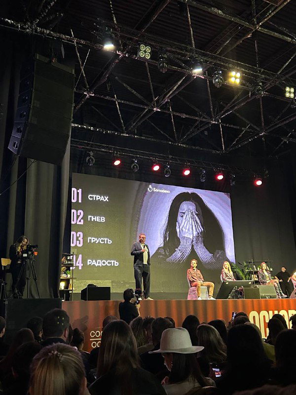 Спикер выступает на сцене «Основатели Show» на фоне слайда с перечислением эмоций: страх, гнев, радость, печаль; зал слушает внимательно.
