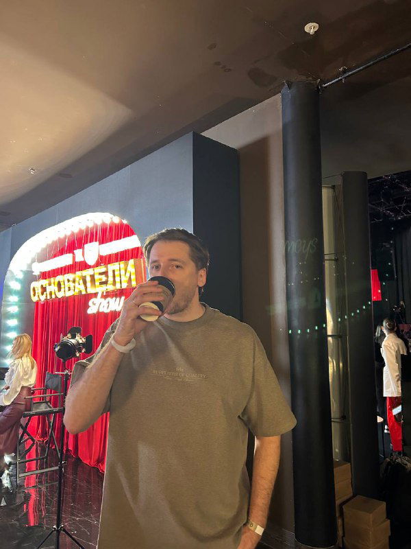 Человек с напитком у внутренней зоны мероприятия на фоне яркой вывески «Основатели Show», неформальная атмосфера и общение гостей.