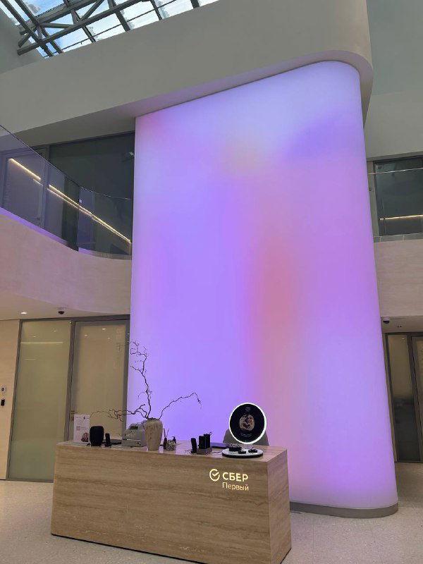 Интерьер лобби или офисного пространства с большой колонной и фиолетовой подсветкой, стойка ресепшн и декоративные элементы.