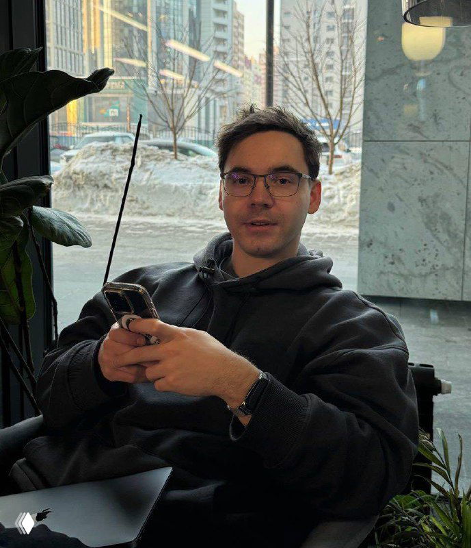 Мужчина в очках и толстовке сидит в кафе у окна с телефоном в руках на фоне зимнего городского пейзажа; иллюстрация к публикации о продвижении в Телеграм.