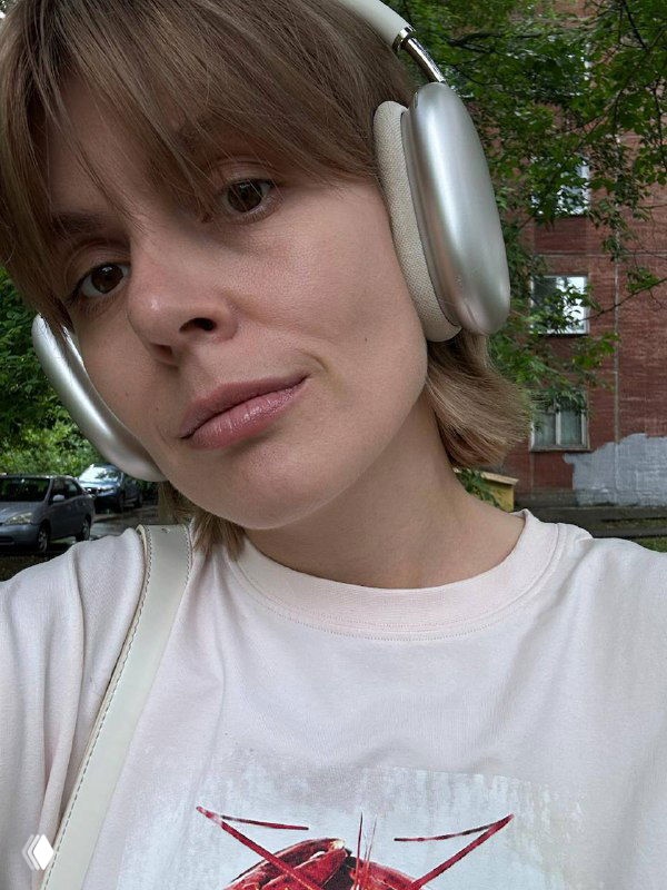 Крупный план молодой женщины в серебристых наушниках на улице, светлая футболка с принтом, зелёные деревья и жилой дом на заднем плане