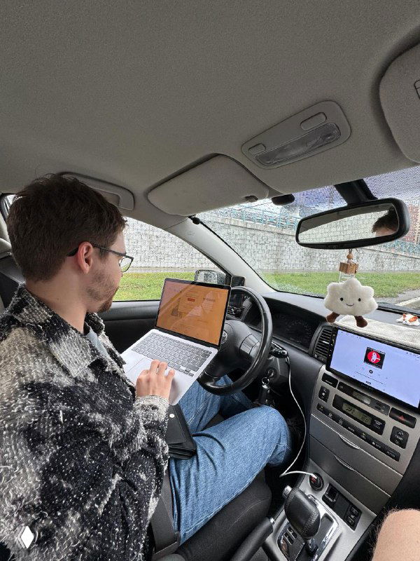 Мужчина в салоне автомобиля с ноутбуком на коленях печатает пост для Telegram; видны руль, торпеда и экран ноутбука, рабочая сцена в машине.