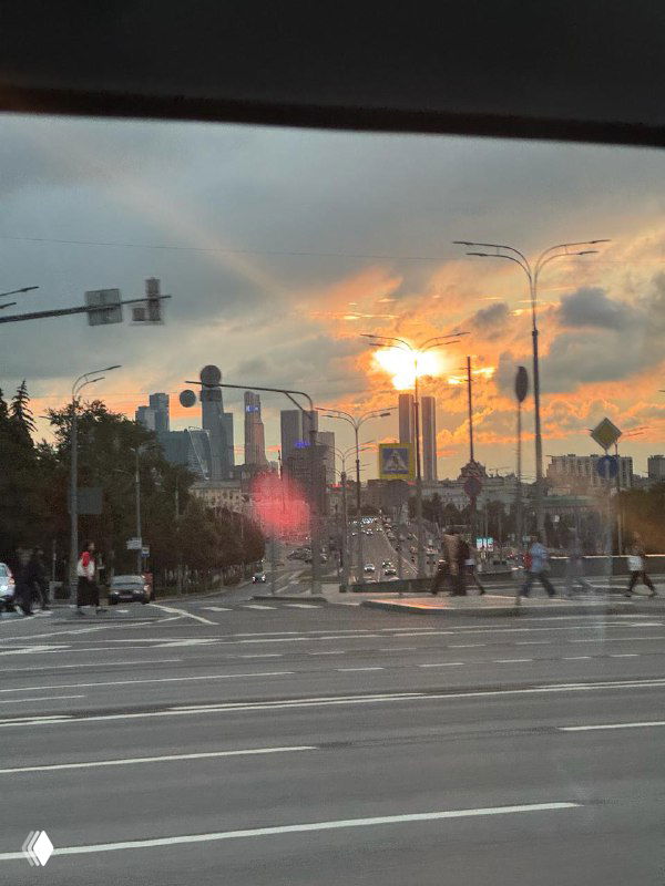 Улица большого города на закате: перекрёсток с машинами и пешеходами, небоскрёбы на фоне солнца, вид через стекло с бликами.