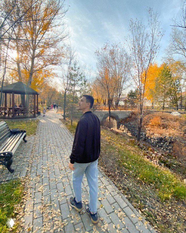 Человек идёт по мощёной аллее в осеннем парке: на заднем плане лавочки, беседка и жёлтая листва — нейтральный кадр со спины, атмосфера прогулки.