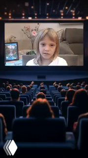 Кадр: школьница Кира на экране монитора во время онлайн-урока; на переднем плане затемнённый зал и силуэты сидящих зрителей, демонстрация прогресса.