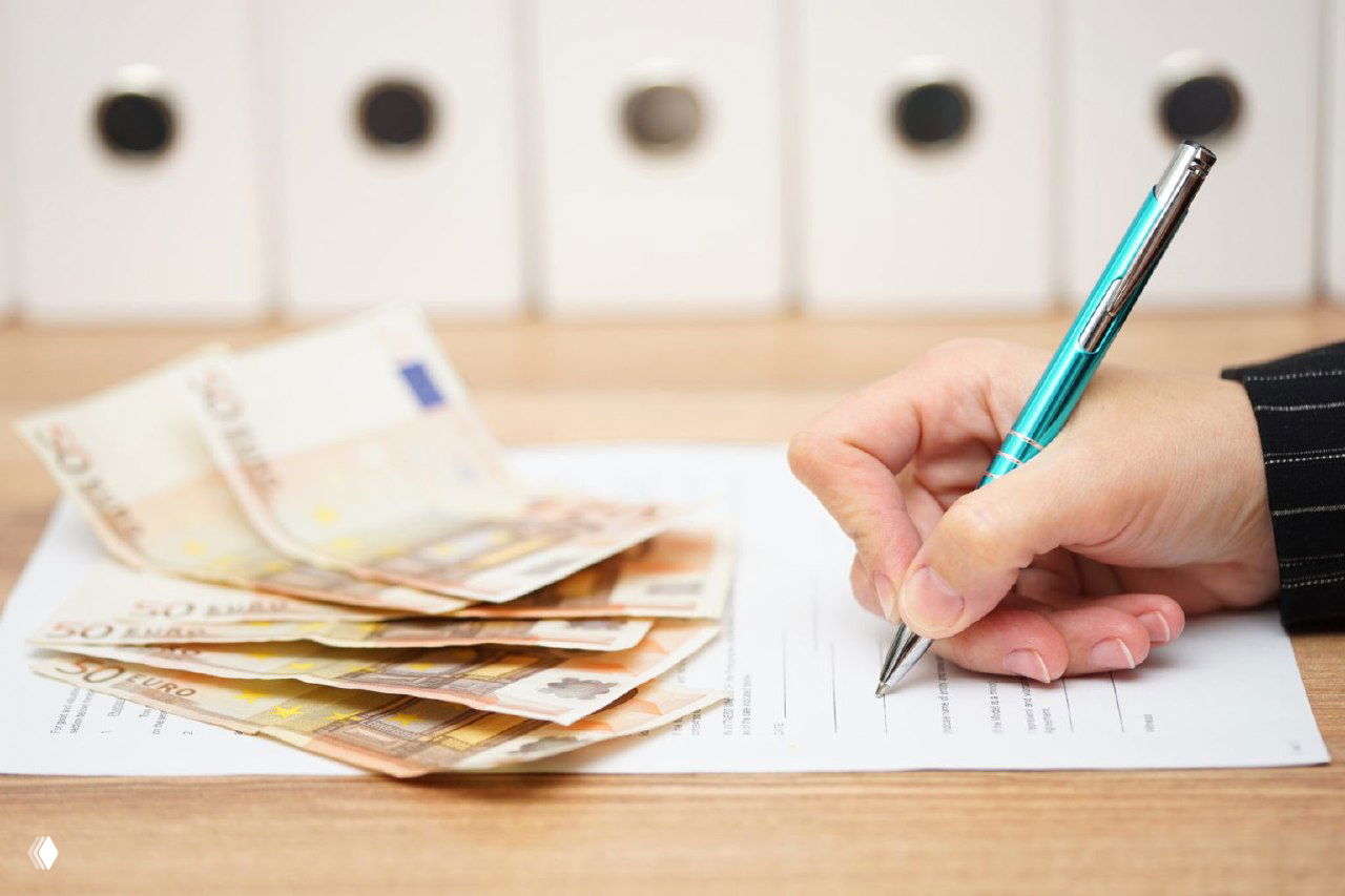 Рука с ручкой подписывает документ на столе рядом с разложенными купюрами евро — изображение иллюстрирует советы по грамотной расписке при оплате недвижимости наличными.