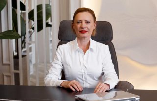 Женщина в белой блузке за рабочим столом в офисе; на столе ноутбук, кресло и фоновые растения, деловой портрет