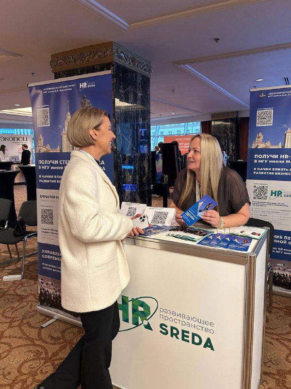 Стенд HR‑SREDA на выставочной зоне саммита: представитель общается с участником у промо‑стола, видны брошюры и брендовые баннеры организации.