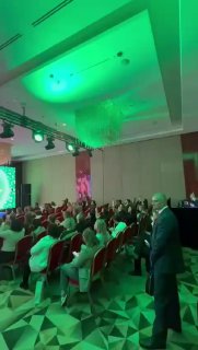 Аудитория конференции во время выступления: ряд кресел с участниками, подсветка сцены зелёным светом и общий вид конференц‑зала.