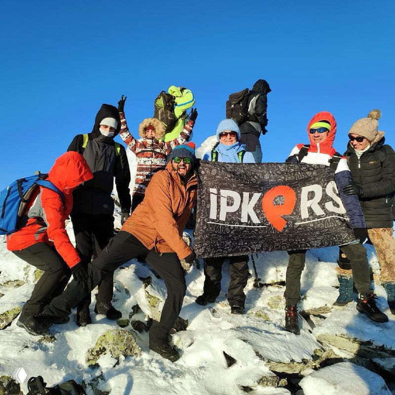 Туристы держат баннер IPKERS на снежной вершине: команда и логотип организатора на фоне голубого неба и снежных камней.