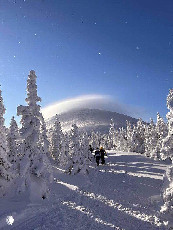 Холмистая вершина в тумане и снегу, два туриста идут по тропе к куполообразной горе под драматичным небом зимнего утра.