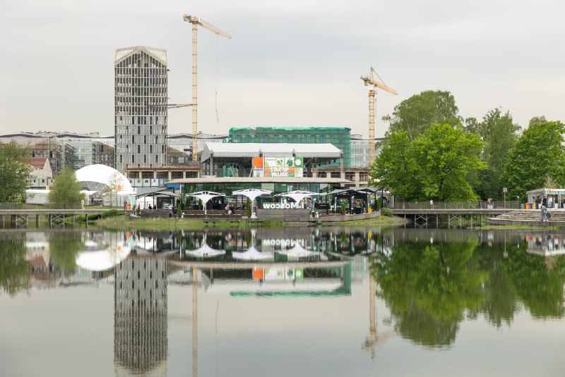 Панорамный вид на площадку Startup Village в Сколково: сцена, временные павильоны, зелёная зона и зеркальное отражение строений и сцены в воде.