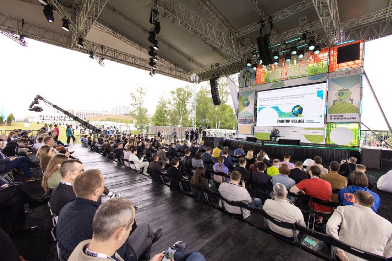 Широкий план аудитории на открытой сцене Startup Village: ряды зрителей, техническое оснащение мероприятия и сцена в перспективе.