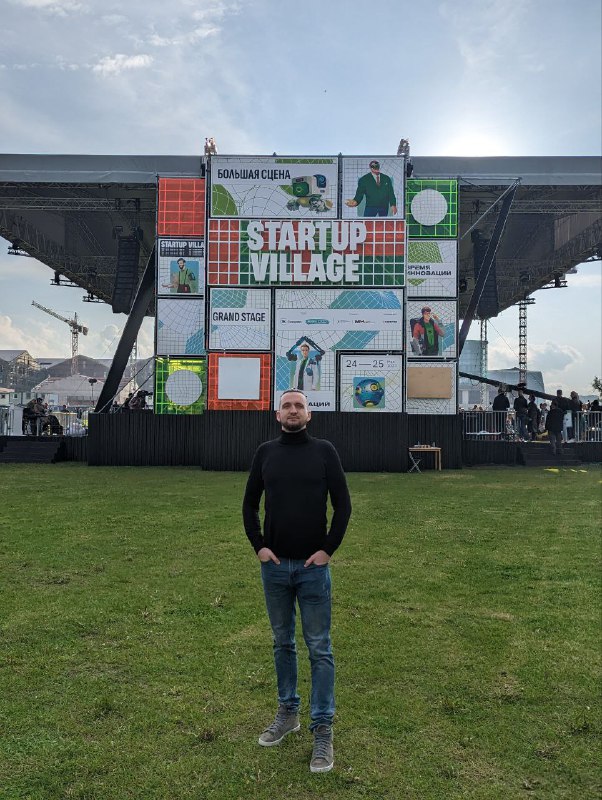 Человек стоит на лужайке перед большой вывеской Startup Village; видна сценическая конструкция, афиши и оформление мероприятия на заднем плане.