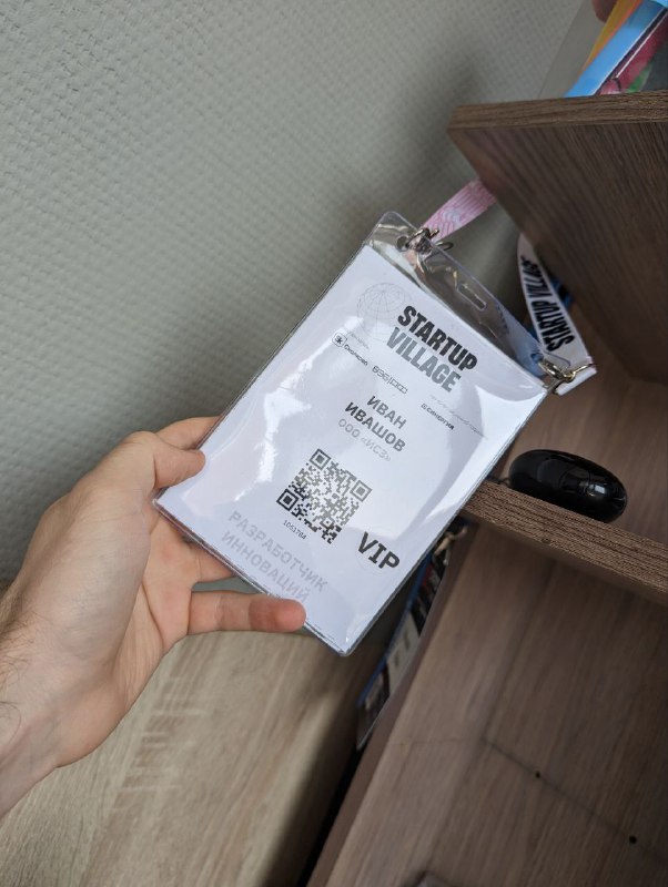 Крупный план бейджа участника Startup Village с текстом, QR-кодом и пометкой статуса, лежащего на столе или поверхности рядом с предметами.
