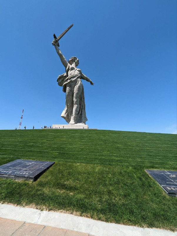 Крупная монументальная скульптура на фоне яркого синего неба, снятая с подножия холма; виден ухоженный зелёный газон и плита у тропинки.