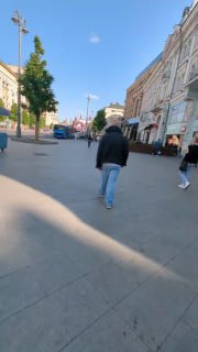 Городская пешеходная улица с прохожими, кадр снят сзади на уровне взрослого человека; видны дома, тротуар и лёгкие тени от деревьев.