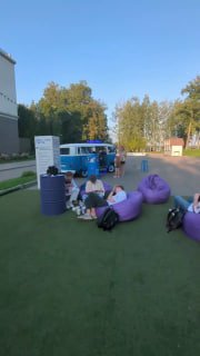 Открытая площадка мероприятия вечером: несколько людей сидят на фиолетовых пуфах на газоне рядом с автомобилем и зданием, атмосферный кадр с общением