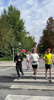 Трое бегунов пересекают пешеходный переход, они в спортивной одежде и масках, вокруг зелёные деревья и городской тротуар под облачным небом.