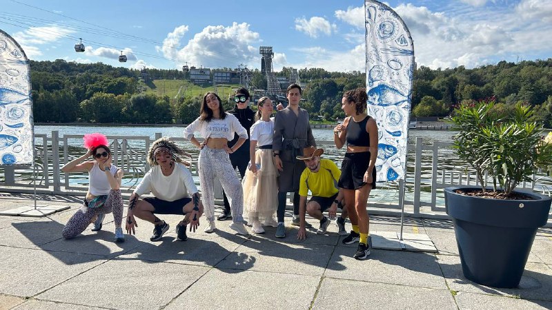 Ещё одна групповая фотография участников забега у набережной: люди в разных костюмах и в повседневной одежде на фоне реки и зелёных склонов.