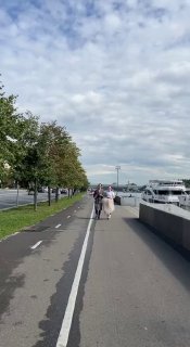 Пешеходно-велосипедная дорожка вдоль реки, на ней прогуливаются люди, видны прогулочные катера и облачное небо над городской набережной.