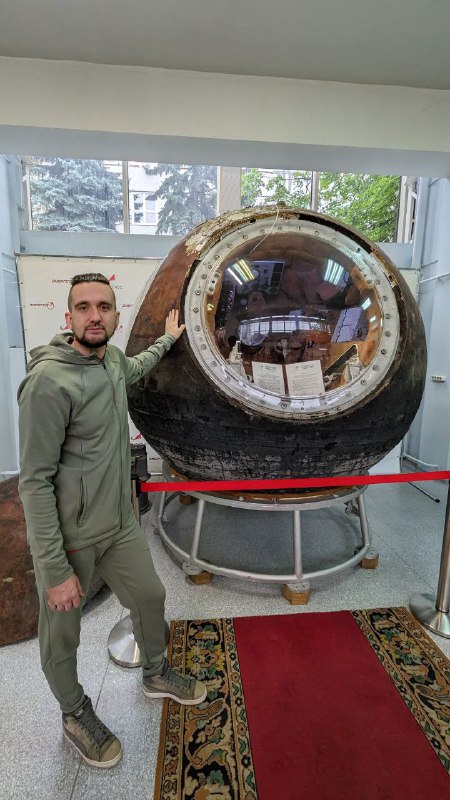 Фотография автора у экспоната: мужчина стоит рядом со спускаемым аппаратом, виден иллюминатор аппарата и ограждение экспозиции внутри зала музея.