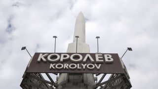 Фотография знака «Королёв» на фоне ракеты и неба с облаками, вид спереди, символичное изображение космонавтики.
