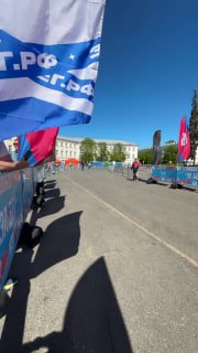 Кадр с флагами, ограждениями и частью линии забега, видна сцена и барьеры у трассы, солнечная погода, волонтёры и организация мероприятия на асфальте.