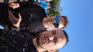 Селфи двух людей в толпе участников на старте или финише, лица крупным планом, многие в синих футболках забега, ощущение праздника и совместного события на солнце.