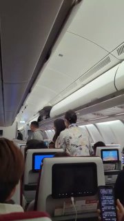 Вид внутри салона самолёта: ряды кресел с встроенными экранами, пассажиры в проходе и закрытые багажные отсеки сверху.