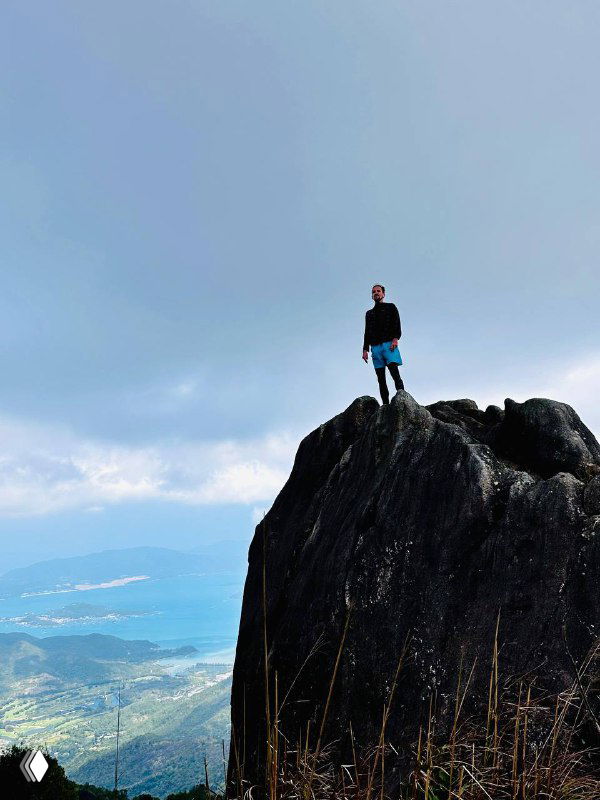 Автор стоит на вершине утеса или утёса в горах, позади открывается панорама побережья и облачное небо — ощущение свободы и высоты.
