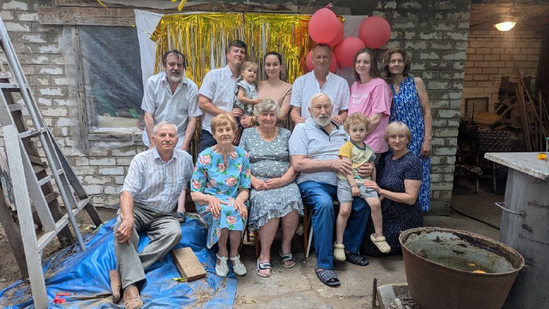Групповое семейное фото на юбилее на фоне украшений и людей разного возраста: бабушка в центре, родственники вокруг.