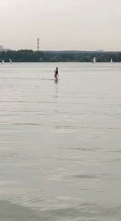 Человек стоит на доске в спокойной воде у далёкого берега, вокруг видны редкие парусные лодки и ровная гладь