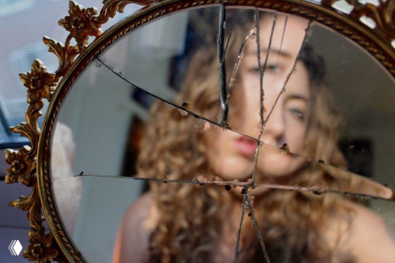 Разбитое старинное зеркало в позолоченной рамке, в осколках видно размытое отражение женщины — метафора раздробленного «я».
