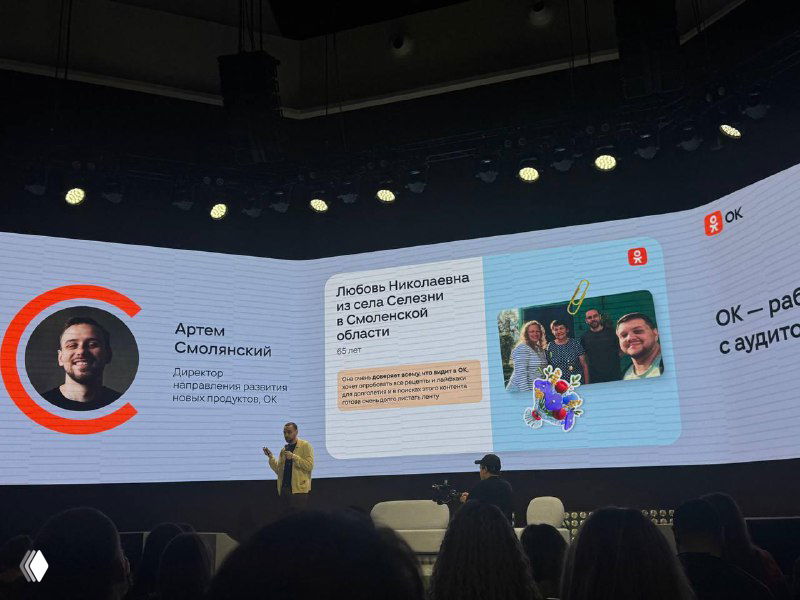 Сцена конференции с крупным экраном, на котором показаны стикеры и примеры пользовательского взаимодействия внутри ОК, присутствуют выступающие и зрители.