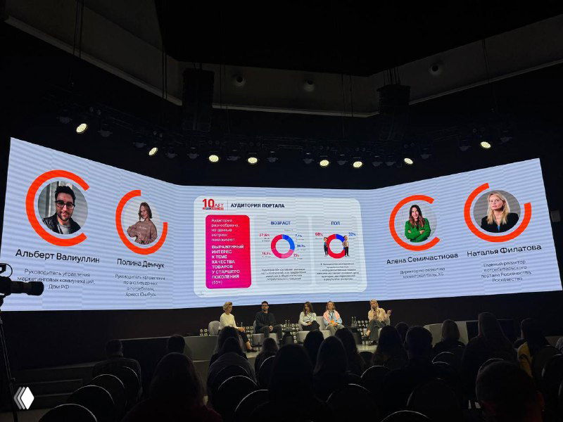 Широкий кадр панели спикеров на сцене с крупным экраном, где видны аватары и блоки с ключевыми данными по аудитории ОК; зал частично заполнен.