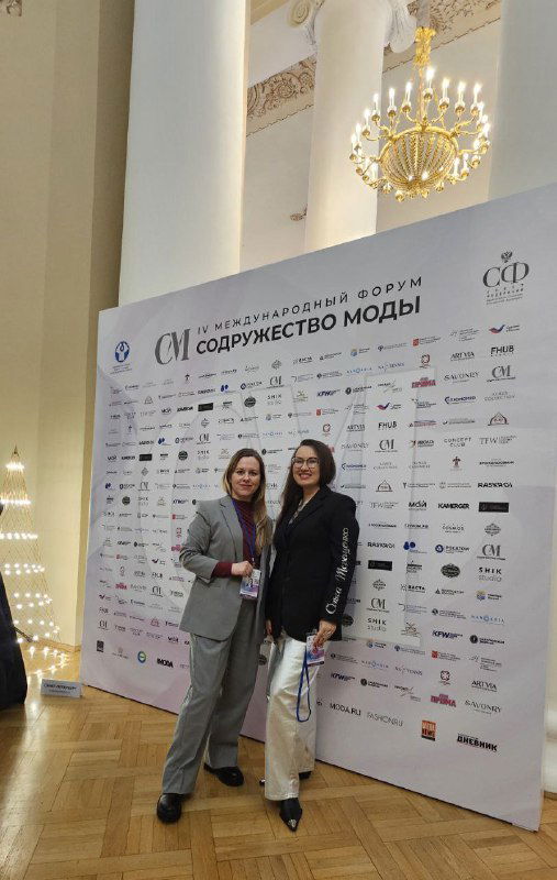 Татьяна Кархалёва и коллега позируют у брендовой фотозоны форума «Содружество моды», на заднем плане логотипы спонсоров и партнёров мероприятия.