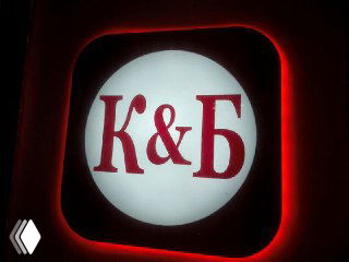 Фотография подсвеченной вывески с буквами «К&Б» на круглой световой панели ночью; изображение иллюстрирует спор вокруг логотипа сети «Красное и Белое».