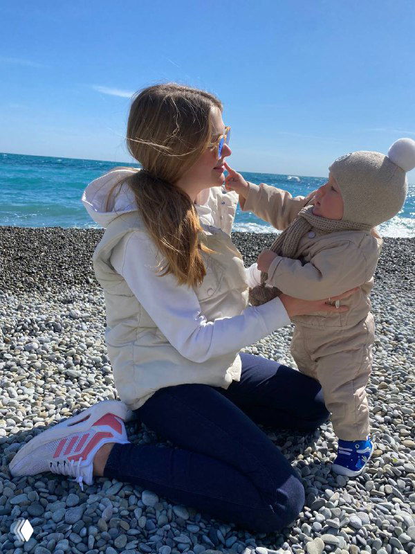 Мама и малыш на галечном пляже: женщина сидит на камнях, держит ребёнка в тёплой одежде с капюшоном, на фоне море и небо.