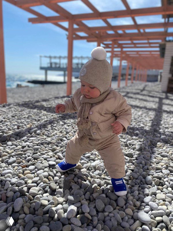 Малыш в тёплом комбинезоне делает первые шаги по гальке у перголы на пляже; видны море и солнечное небо, семейная прогулка.