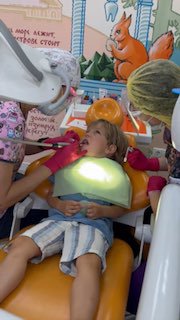 Ребёнок в стоматологическом кресле во время профессиональной чистки молочных зубиков; врач в перчатках, яркая детская декорация